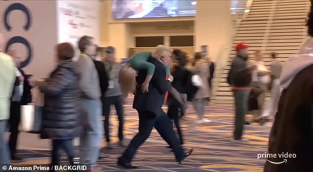 A still from the Borat film series shows Cohen (in Trump costume, center) running over the shoulder with an actress through a rally venue.