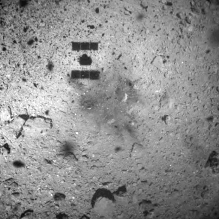 The shadow of the unmanned probe Hayabusa 2 glides over the surface of the distant asteroid Ryugu, shortly before taking a bite of sand and dust there.  This bite has now been analyzed by scientists.  Image access point